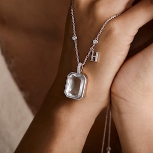 May include: A silver-toned necklace featuring a large, rectangular, clear faceted pendant in a metallic setting. The chain includes two small round bezel-set stones and a dangling letter H charm embellished with tiny clear stones along its surface.