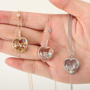 May include: Three heart-shaped locket necklaces featuring clear glass fronts and metallic frames in gold, rose gold, and silver tones. Each locket contains small decorative charms inside, suspended from matching delicate link chains with a single crystal accent.