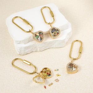 May include: Three gold-toned heart-shaped lockets attached to oval key rings. The lockets feature clear glass fronts framed with small crystals. One locket is open, revealing small colorful charms inside, with additional charms scattered nearby.