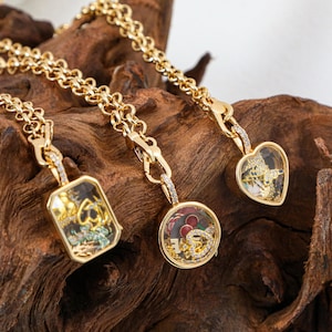 May include: Three gold necklaces with pendant charms. One pendant is rectangular, one is round, and one is heart-shaped. Each charm contains decorative elements and is encrusted with small, clear gemstones. The chains are a thick, linked design.
