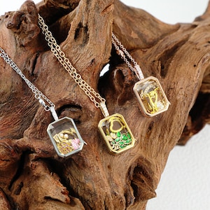 May include: Three rectangular pendant necklaces with delicate chains. One is silver, one gold, and one rose gold. Each pendant has a clear, rectangular frame with colorful interior designs, including gems and floral elements. The necklaces rest on a piece of dark wood.