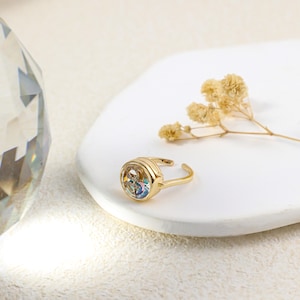 May include: A gold-colored adjustable ring with a round, iridescent stone set in a bezel. The ring is on a white surface with dried flowers. The ring's band has a simple, elegant design.