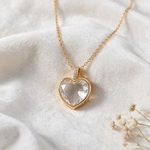 Peut inclure: Un collier doré avec un pendentif en forme de cœur en pierre transparente à facettes, serti dans une monture en or poli. Le pendentif est suspendu à une fine chaîne dorée, avec une bélière ornée de petites pierres claires sur le dessus.