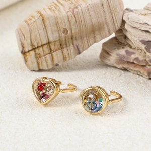 May include: Two gold-toned adjustable rings. One ring features a heart-shaped setting containing small red and clear stones. The second ring has a circular setting with a gold-toned anchor symbol encrusted with clear stones over colourful accents.