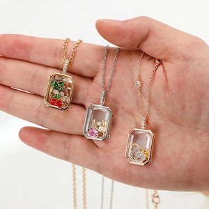 Peut inclure: Trois colliers avec pendentifs rectangulaires et façades en verre transparent. Le premier présente des breloques cerises rouges, le deuxième contient une pierre rose et des perles, et le troisième un cœur ailé. Chaque pendentif est sur une chaîne.