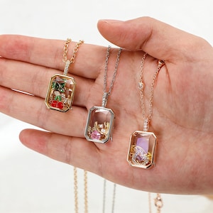 May include: Three rectangular pendant necklaces with delicate chains. One is gold-toned with a cherry design, another silver-toned with pink and pearl accents, and the third rose gold-toned with a purple gem. Each pendant has a clear, glass-like center.