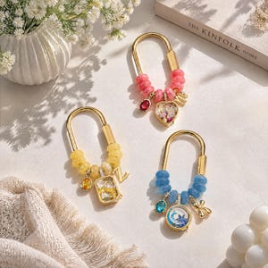 Peut inclure: Trois porte-clés mousquetons dorés en forme de U avec des perles colorées roses, jaunes et bleues. Chaque porte-clés comprend une breloque suspendue, une lettre et une petite pierre précieuse ovale. Les breloques contiennent des éléments décoratifs.