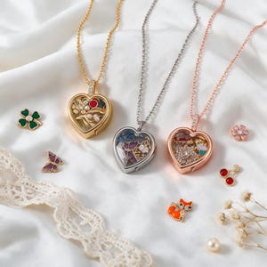 May include: Three heart-shaped locket necklaces in gold, silver, and rose gold tones, each containing small charms. Scattered around are individual charms including a green clover, a purple butterfly, a pink flower, red cherries, and an orange fox.