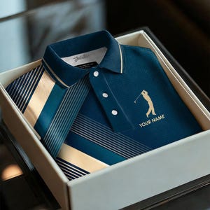以下が含まれることがあります： 金色のゴルファーのシルエットと「YOUR NAME」の文字が入ったネイビーブルーのゴルフ用ポロシャツが、白いギフトボックスに入っています。シャツはテクスチャード加工で、襟と袖にはストライプのデザインが施されています。「Jubilee」のブランド名が見えます。