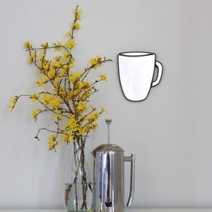 May include: A white ceramic coffee mug with a black outline hangs on a white wall. A vase with yellow flowers sits on a white surface next to a silver french press.