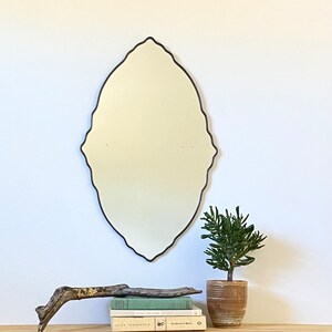 May include: A decorative oval mirror with a black frame and a scalloped edge. The mirror is hanging on a white wall above a wooden shelf with books and a potted plant.