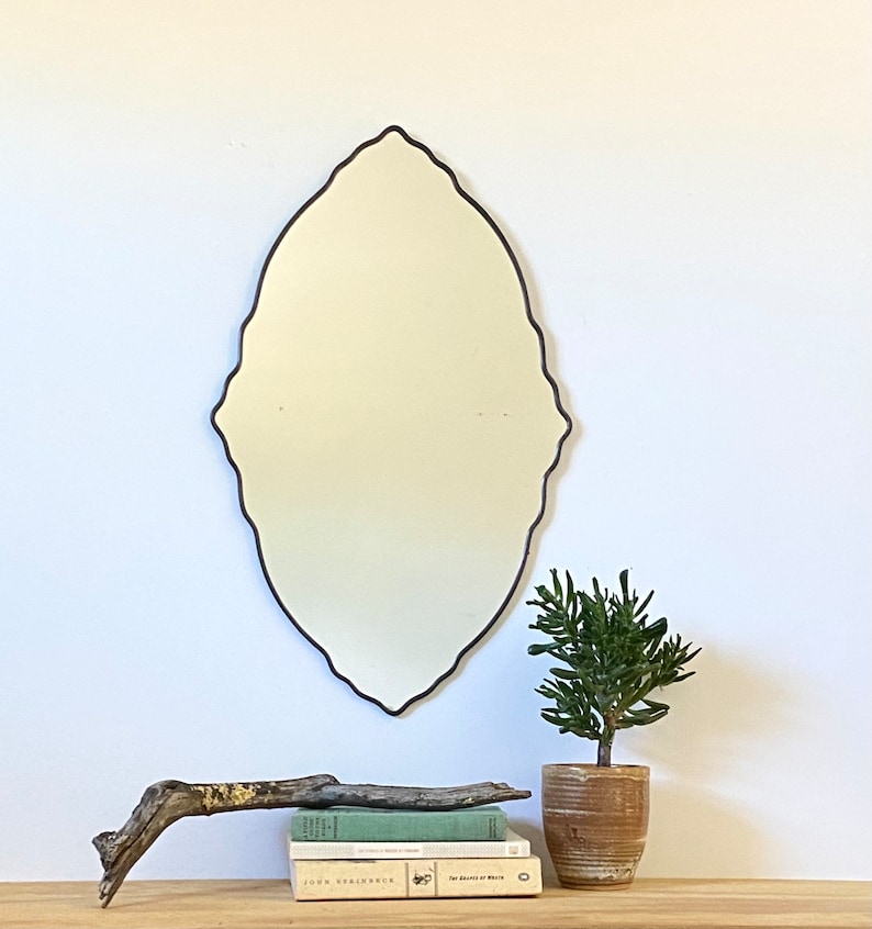 May include: A decorative oval mirror with a black frame and a scalloped edge. The mirror is hanging on a white wall above a wooden shelf with books and a potted plant.