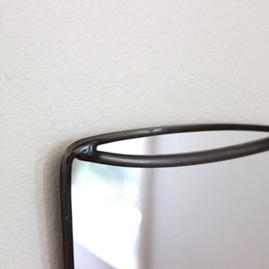 May include: A black metal framed mirror shaped like a coffee cup. The mirror is hanging on a white wall.