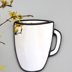 May include: A white mug-shaped mirror with a black outline. The mirror is hanging on a light gray wall with yellow flowers in the background.