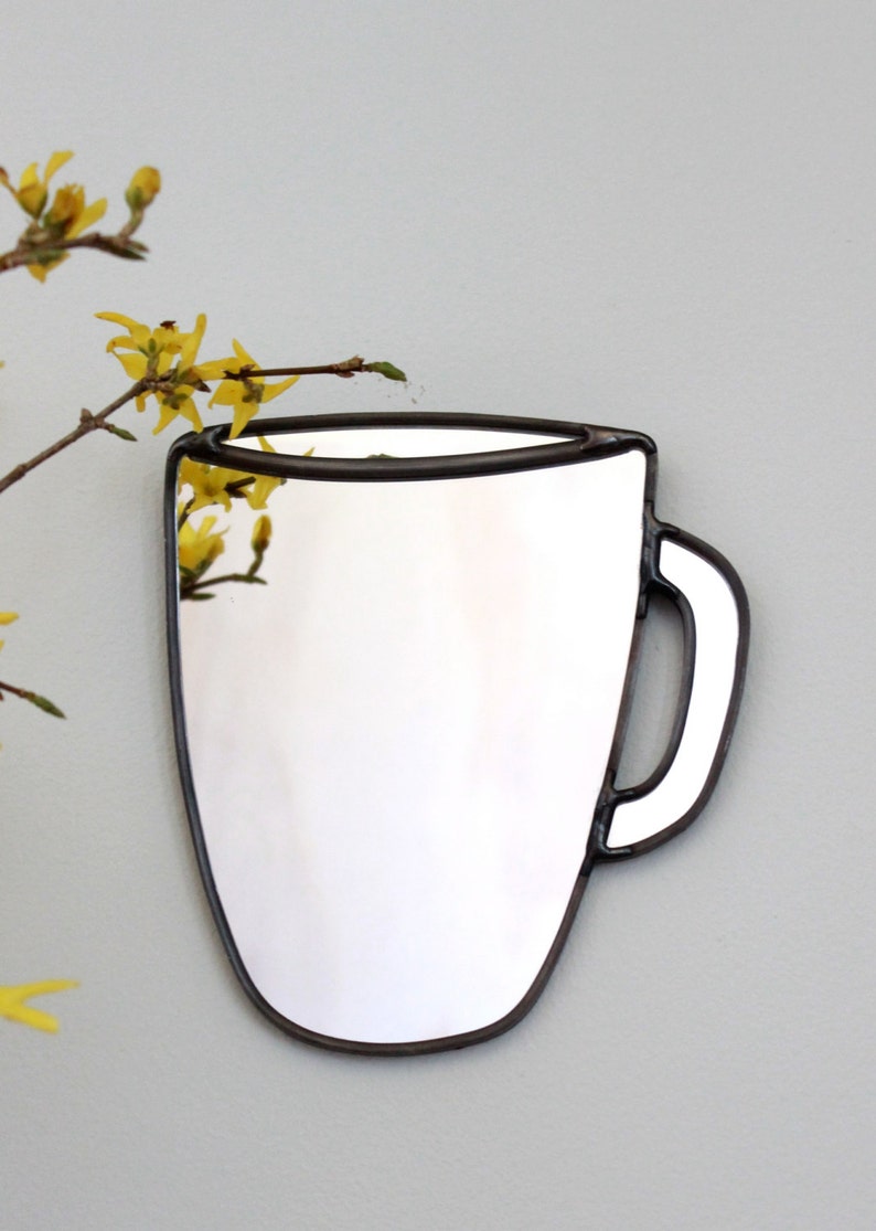 May include: A white mug-shaped mirror with a black outline. The mirror is hanging on a light gray wall with yellow flowers in the background.