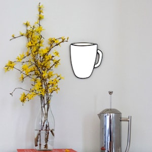 May include: A white ceramic coffee mug with a black outline hangs on a white wall. A vase with yellow flowers sits on a white surface next to a silver French press and a red book with the text "DESIGN*SPONGE at HOME".