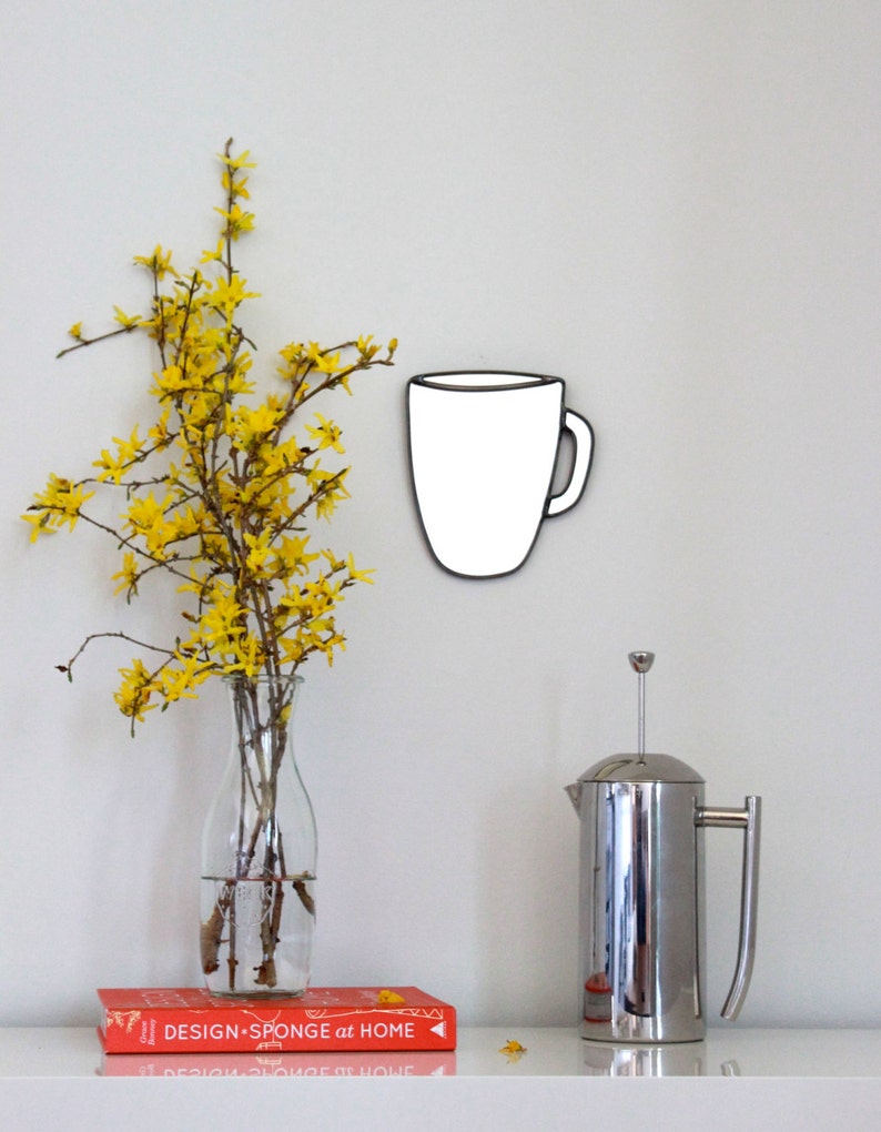 May include: A white ceramic coffee mug with a black outline hangs on a white wall. A vase with yellow flowers sits on a white surface next to a silver French press and a red book with the text "DESIGN*SPONGE at HOME".