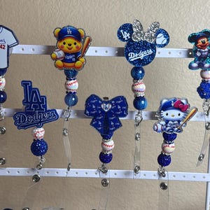 May include: A collection of retractable badge holders featuring various designs. The designs include a baseball jersey, a bear, Minnie Mouse, Mickey Mouse, a bow, Hello Kitty, and the Dodgers logo. Each badge holder has a beaded accent with baseball and blue beads.