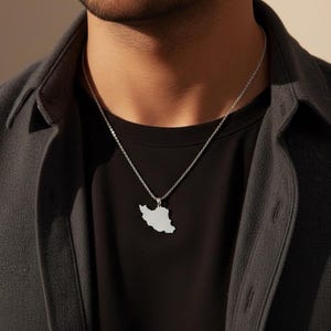 May include: A silver-colored pendant necklace featuring the outline of a country. The pendant hangs from a delicate chain. The necklace is worn over a black t-shirt and a dark gray jacket.