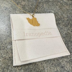 Peut inclure: Un collier dor&eacute; avec un pendentif en forme de la carte de l'Iran. Le collier est pos&eacute; sur une pochette cr&egrave;me brod&eacute;e du mot "Iranopedia". La pochette est pli&eacute;e et poss&egrave;de un rabat.