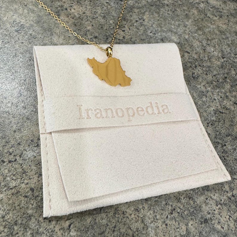 Peut inclure: Un collier dor&eacute; avec un pendentif en forme de la carte de l'Iran. Le collier est pos&eacute; sur une pochette cr&egrave;me brod&eacute;e du mot "Iranopedia". La pochette est pli&eacute;e et poss&egrave;de un rabat.