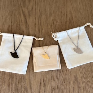 Peut inclure: Trois colliers avec des pendentifs en forme de carte d'un pays. L'un est noir, l'autre dor&eacute; et le dernier argent&eacute;. Chaque collier est pr&eacute;sent&eacute; dans un petit sac ou une pochette &agrave; cordon de serrage blanc cass&eacute;, sur fond de bois.