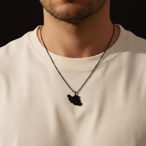 Puede incluir: Un collar con colgante negro que representa el contorno de un país. El colgante cuelga de una fina cadena. El collar se lleva sobre una camiseta blanca.