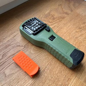 Puede incluir: Un dispositivo repelente de mosquitos verde con una rejilla negra en la parte delantera y un bot&oacute;n negro. El dispositivo est&aacute; etiquetado como "Mosquito Repellent". Tambi&eacute;n se muestra un peque&ntilde;o clip naranja.