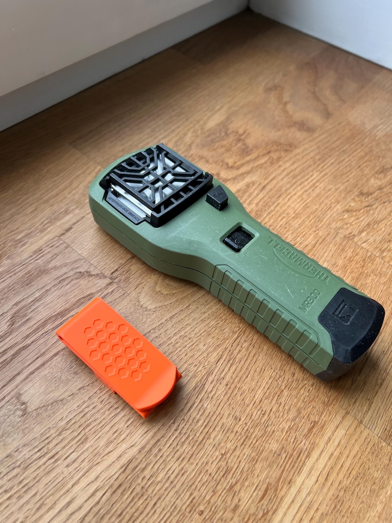 Puede incluir: Un dispositivo repelente de mosquitos verde con una rejilla negra en la parte delantera y un bot&oacute;n negro. El dispositivo est&aacute; etiquetado como "Mosquito Repellent". Tambi&eacute;n se muestra un peque&ntilde;o clip naranja.