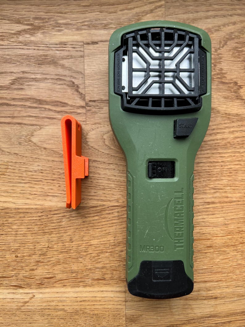 Puede incluir: Dispositivo repelente de mosquitos Thermacell MR300 verde con un bot&oacute;n de inicio negro y una rejilla negra. El dispositivo tiene un clip de cintur&oacute;n negro y naranja.