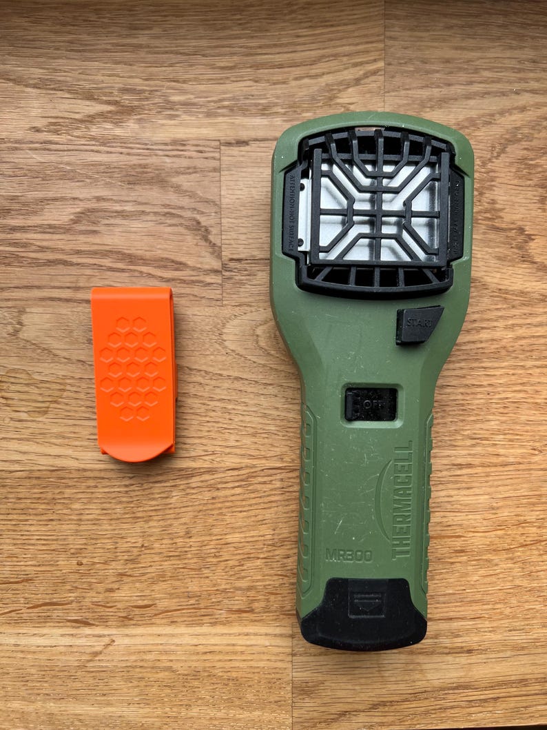 Puede incluir: Un dispositivo repelente de mosquitos Thermacell MR300 verde con un clip negro y un filtro blanco. El dispositivo tiene un bot&oacute;n negro etiquetado como "Start" y un bot&oacute;n negro etiquetado como "Off".
