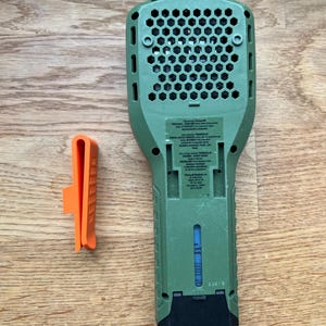 Puede incluir: Dispositivo repelente de insectos verde y negro con un indicador de combustible transparente. El dispositivo tiene una rejilla en forma de panal en la parte posterior y una etiqueta con instrucciones de seguridad. El dispositivo viene acompa&ntilde;ado de un peque&ntilde;o clip naranja.