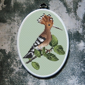 May include: Oval cross-stitch embroidery of a hoopoe bird perched on a branch with green leaves. The bird has brown, black, and white markings, a long beak, and a distinctive crest. The background is a light green fabric.