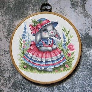 May include: Cross-stitch embroidery of a gray bunny wearing a blue and pink dress and hat, surrounded by flowers and greenery. The embroidery is set in a wooden hoop with a metal clasp.