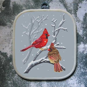 Puede incluir: Punto de cruz de dos cardenales posados en una rama nevada. El cardenal macho es rojo brillante, mientras que la hembra es marrón y roja. El fondo es azul claro con nieve cayendo, enmarcado en un cuadrado.