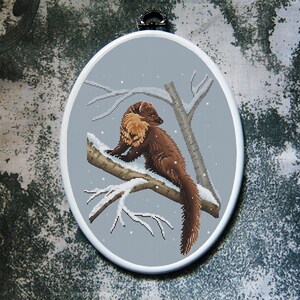 May include: Oval cross-stitch embroidery depicting a brown pine marten on a snow-covered branch. The marten has a light brown chest and is set against a grey background with falling snow. The embroidery is framed in a white oval.