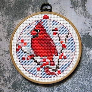 Könnte beinhalten: Kreuzstich-Stickerei eines roten Kardinals auf einem Ast mit roten Beeren. Das Bild ist in einem Holzrahmen mit blau-weißem Hintergrund gefasst. Der Rahmen hat eine Metallschlaufe zum Aufhängen.