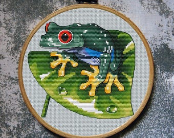 May include: A cross-stitch embroidery of a vibrant green tree frog with red eyes, perched on a large green leaf. The frog has yellow feet and is set against a light gray background, framed in a wooden hoop.
