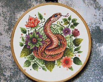 May include: Cross-stitch embroidery of a snake with a red and orange pattern, blue eyes, and a forked tongue, surrounded by green leaves, purple and orange flowers, and a wooden hoop. The hoop has a metal ring for hanging.