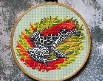 May include: Cross-stitch embroidery of a poison dart frog in a wooden hoop. The frog has a red head and black and white patterned body, resting on a red and yellow leaf. The background is a light green fabric.