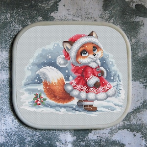 May include: Cross-stitch image of a fox wearing a red winter coat and hat, set in a snowy scene. The fox has orange fur, brown boots, and a white-tipped tail. The background shows a snowy hill and some small trees.