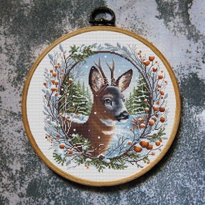 May include: A cross-stitch embroidery of a deer in a winter scene, set in a wooden hoop. The deer is brown with antlers, surrounded by snow-covered trees and orange berries. The background is a snowy landscape.