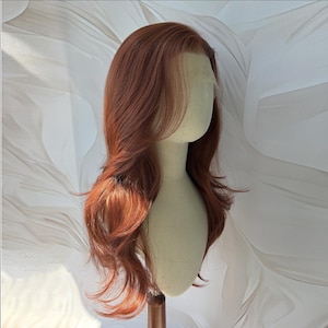 May include: A long, wavy wig in a vibrant copper-red shade, styled with a side part and soft layers. The wig features a sheer lace front hairline and is displayed on a neutral-toned mannequin head, showcasing a natural-looking hair texture.