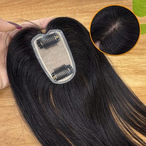 Puede incluir: Un postizo de cabello negro con base de encaje y dos clips para una sujeción segura. La pieza está diseñada para añadir volumen y cobertura, mezclándose a la perfección con el cabello natural. La imagen también muestra un primer plano de la raíz del cabello.