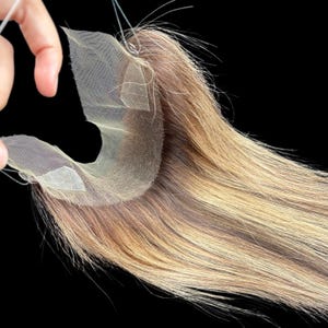 Puede incluir: Primer plano de una pieza de cabello con base de encaje y mechones de cabello largos, lisos y castaños claros. La pieza de cabello se sostiene parcialmente, revelando la fina malla del encaje. El color del cabello pasa de raíces más oscuras a puntas más claras.