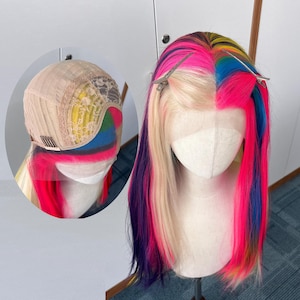 May include: A brightly coloured wig with a split design, featuring vibrant pink, blue, yellow, and purple strands. The wig is styled on a mannequin head, showcasing a lace front and a detailed cap construction. Hair clips secure the wig.