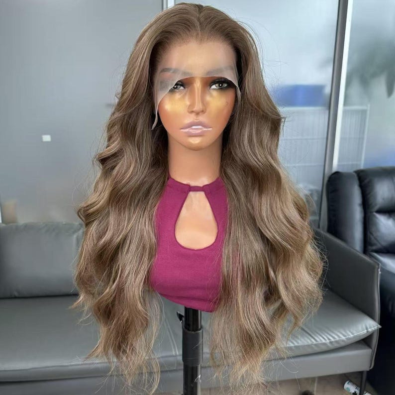 May include: A long, wavy wig in shades of light brown and blonde, styled on a mannequin head. The wig features a lace front and a natural-looking hairline. The mannequin is wearing a maroon top with a keyhole neckline.