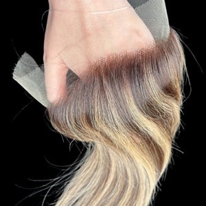 Può includere: Una parrucca frontale in pizzo con una miscela di capelli castani, biondi e castano chiaro. I capelli sono acconciati in onde sciolte e il pizzo è di colore beige chiaro. La parrucca è tenuta su uno sfondo nero.