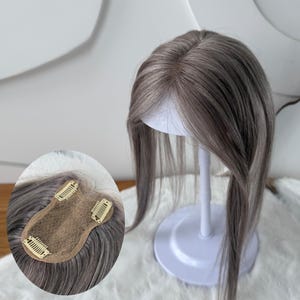 May include: A hair topper with straight, ash-toned brown and gray hair strands. The piece features a central part and is displayed on a white stand. An inset shows the underside with a tan mesh base and three metal pressure-sensitive clips.