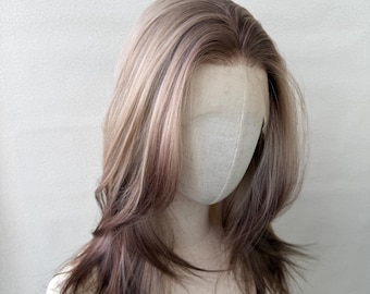May include: A shoulder-length wig featuring a layered cut with a center part. The hair color is a blend of light ash blonde at the roots transitioning into soft mauve and dusty rose pink tones throughout the mid-lengths and the ends.